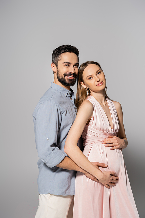 Smiling bearded man hugging elegant pregnant woman in pink dress and looking at camera together on grey background, expecting parents concept, husband and wife