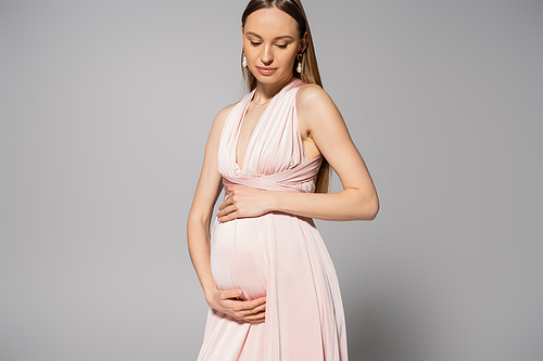 Elegant and fair haired expecting mother in pink dress touching belly and looking down while standing on grey background, maternity fashion concept, fashionable pregnancy attire