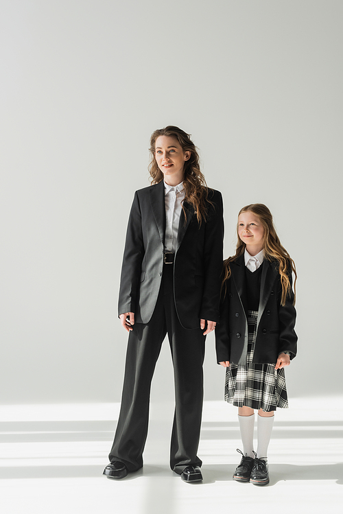 working mother and schoolgirl, cheerful girl in school uniform standing with businesswoman in suit on grey background, holding hands, formal attire, fashionable family, bonding, modern parenting