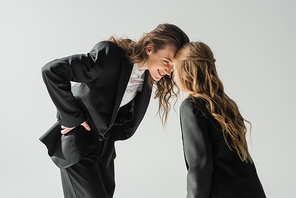 happy mother and daughter having fun, woman in suit and schoolgirl in uniform looking at each other and smiling on grey background in studio, fashionable family, formal attire