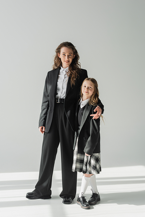 modern mother and daughter, businesswoman in suit hugging schoolgirl in uniform with plaid skirt, on grey background, blazers, getting ready for new school year, looking at camera, formal attire