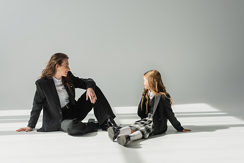 fashionable mother and daughter, businesswoman in suit sitting and looking at schoolgirl in uniform with plaid skirt on grey background, blazers, new school year, looking at each other