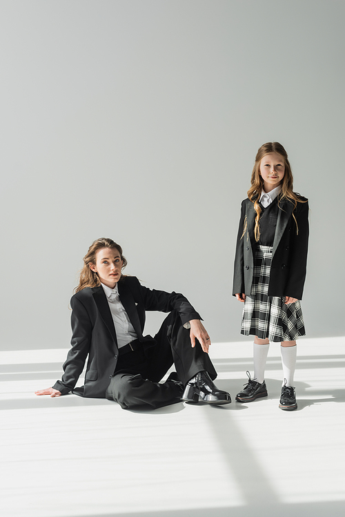 working mother and child, businesswoman in suit sitting near schoolgirl in uniform with plaid skirt on grey background, blazers, new school year, looking at camera, formal attire