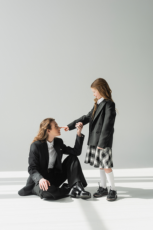 schoolgirl touching nose of working mother, cheerful girl in school uniform looking at businesswoman in suit on grey background, formal attire, fashionable family, bonding, modern parenting