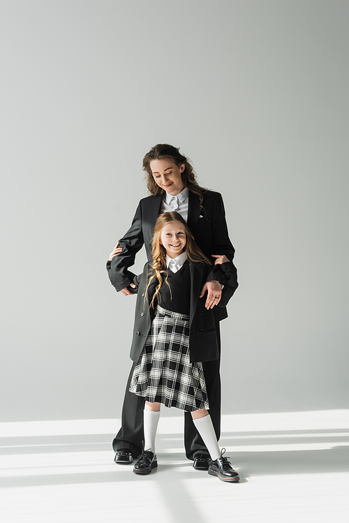 modern parenting, fashionable businesswoman in suit hugging daughter in school uniform with plaid skirt and standing together on grey background, happy mother and child