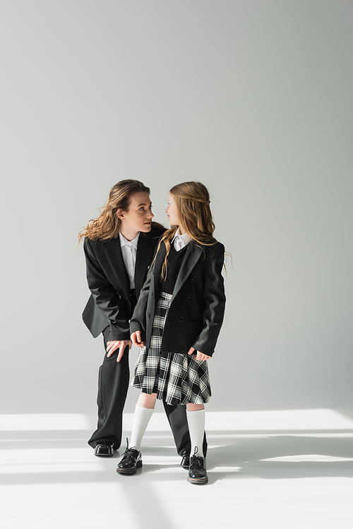 schoolgirl looking at working mother, cheerful girl in school uniform standing with businesswoman in suit on grey background, formal attire, fashionable family, bonding, modern parenting