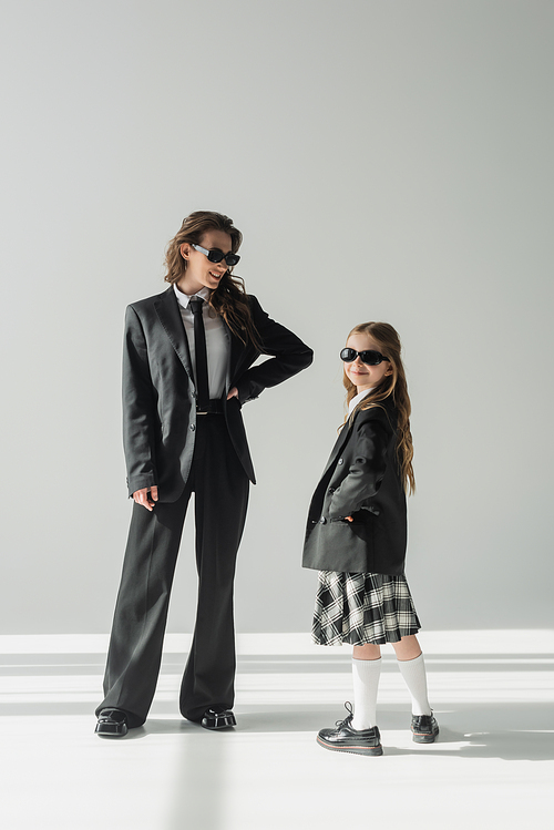 cheerful woman with her daughter, businesswoman in suit posing with hand on hip and schoolgirl in sunglasses and uniform standing together on grey background in studio, formal attire