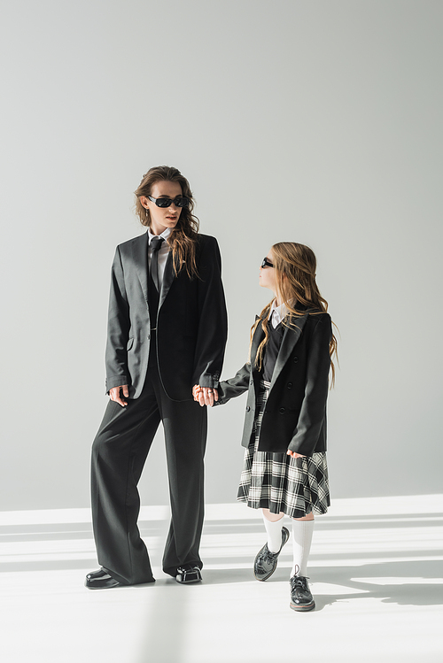 stylish mother and child holding hands, businesswoman in formal attire and sunglasses looking at schoolgirl in uniform on grey background in studio, fashionable family