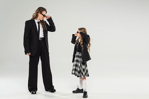 stylish mother and child wearing sunglasses, happy businesswoman in formal attire looking at schoolgirl in uniform on grey background in studio, fashionable family, modern parenting