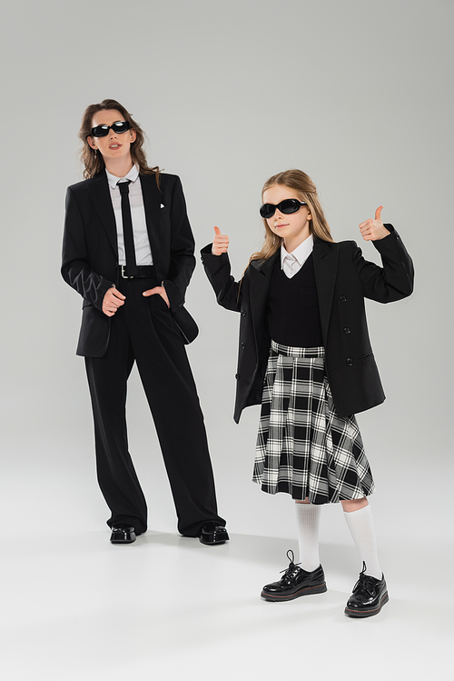 stylish schoolgirl in sunglasses and uniform showing thumbs up and standing near modern mother on grey blurred background in studio, formal attire, back to school