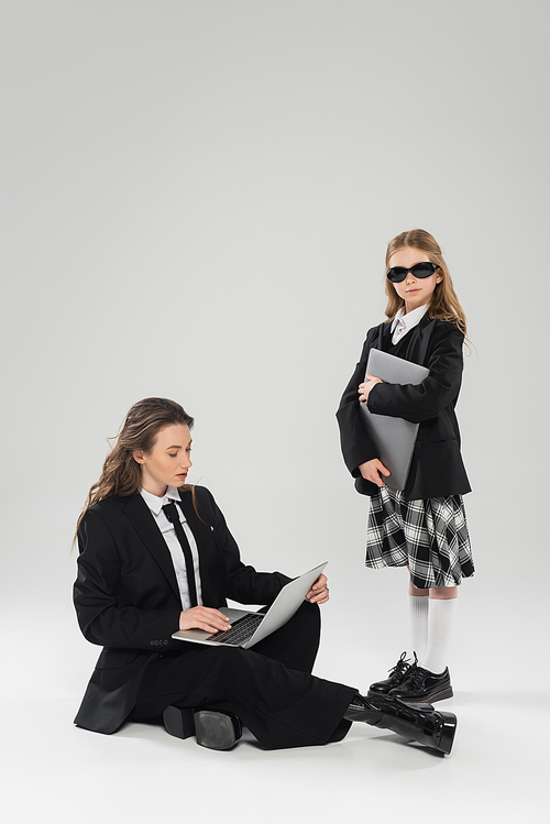 digital nomadism, fashionable woman in suit using laptop near daughter in school uniform and sunglasses on grey background, remote work, working mother, business attire