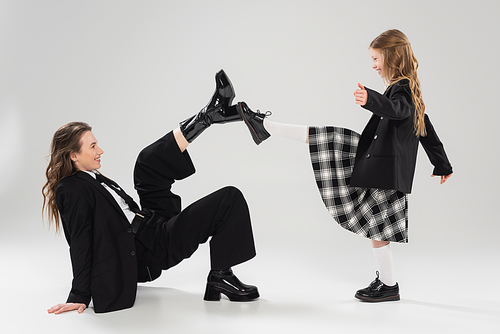 modern parenting, having fun, side view of happy mother in suit and child in school uniform touching boots of each other on grey background in studio, blazer, businesswoman, back to school