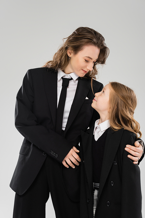 modern parenting, cheerful businesswoman in suit hugging daughter in school uniform and standing together on grey background, happy mother and child, back to school