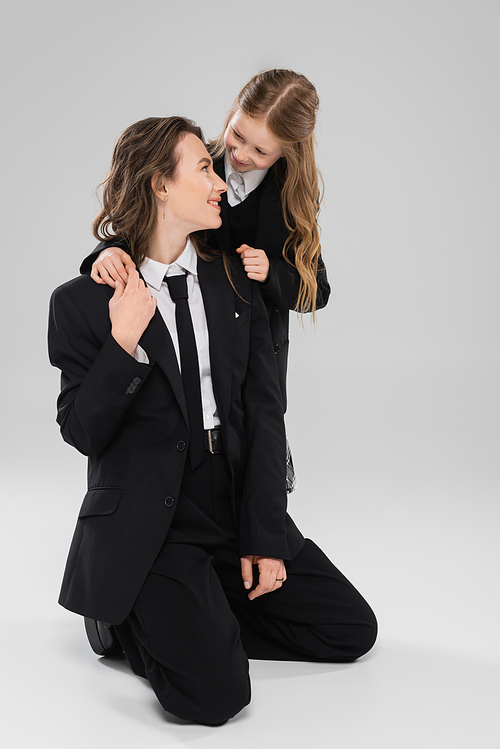 happy schoolgirl hugging working mother, cheerful girl in school uniform looking at mom in suit on grey background, formal attire, fashionable family, bonding, modern parenting