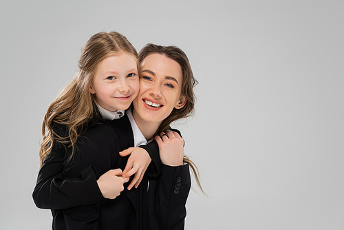 smiling schoolgirl hugging mother in suit, girl in school uniform and her mom in business attire on grey background, fashionable family, bonding, modern parenting, back to school