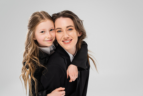 cheerful schoolgirl hugging mother in suit, girl in school uniform and her mom in business attire on grey background, fashionable family, bonding, modern parenting, back to school, portrait