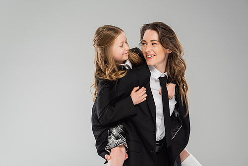 modern parenting, happy mother piggybacking her daughter, working mom in business attire and schoolgirl in uniform on grey background in studio, fashionable family, having fun