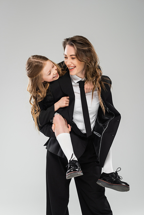 mother and daughter having fun, cheerful woman piggybacking schoolgirl, working mom in formal attire and kid in uniform on grey background in studio, modern parenting