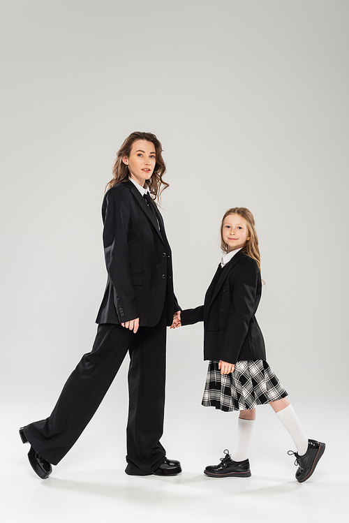 mother and schoolgirl holding hands, woman in business attire and happy girl in school uniform standing together on grey background, modern parenting, back to school