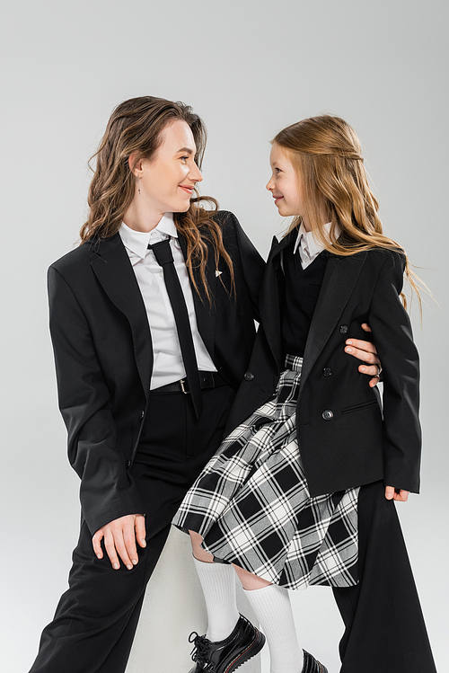 working mother and daughter, happy businesswoman sitting on concrete stool near kid in uniform on grey background in studio, formal attire, modern parenting, fashion shoot