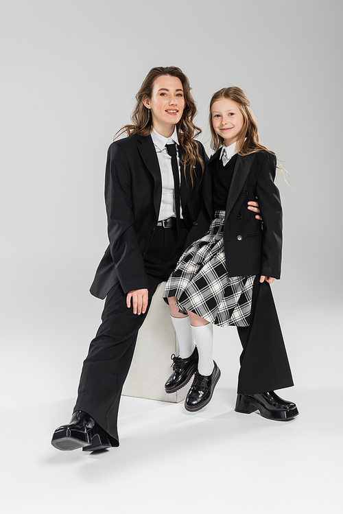 working mother and daughter, happy businesswoman and schoolgirl sitting on concrete stool together on grey background in studio, formal attire, modern parenting, fashion shoot