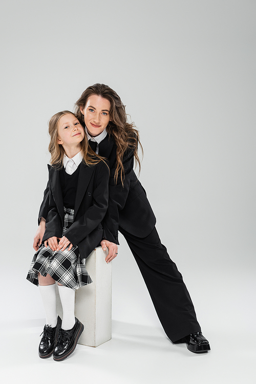 mother and child, happy businesswoman in formal attire standing near schoolgirl sitting on concrete stool on grey background in studio, modern parenting, fashion shoot