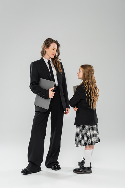 working mother and daughter, digital nomadism, remote work, e learning, businesswoman in suit and schoolgirl standing together with laptops on grey background in studio, modern parenting