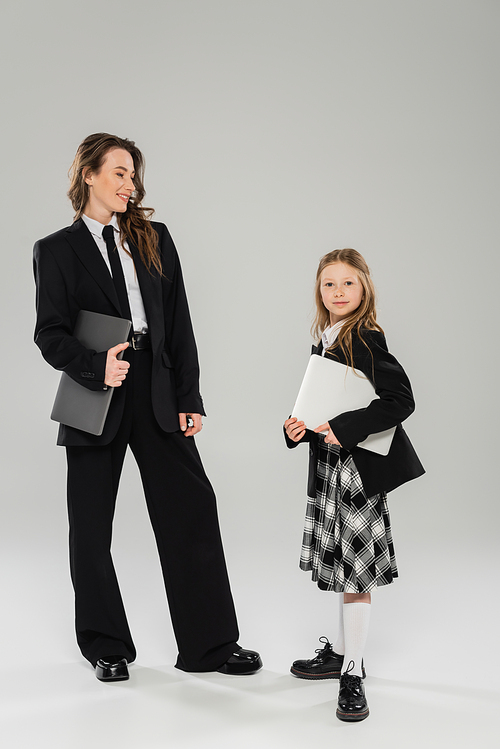 mother and daughter, digital nomadism, remote work, e learning, happy businesswoman in suit and schoolgirl standing together with laptops on grey background in studio, modern parenting