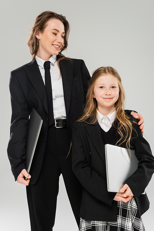 mother and daughter, digital nomadism, remote work, e learning, businesswoman in suit and girl standing together with laptops on grey background in studio, bonding, modern parenting