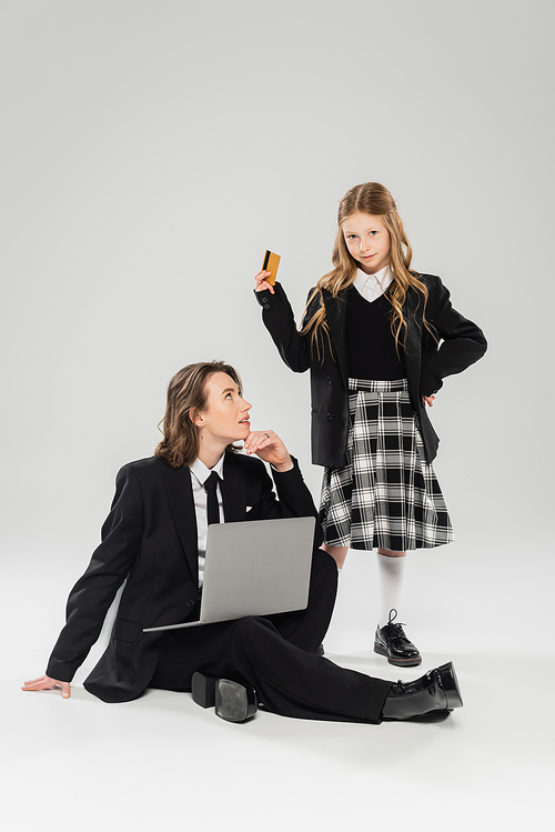 mother and schoolgirl, digital nomadism, girl holding credit card near working mom using laptop on grey background in studio, modern parenting, business attire, online purchase, e-commerce