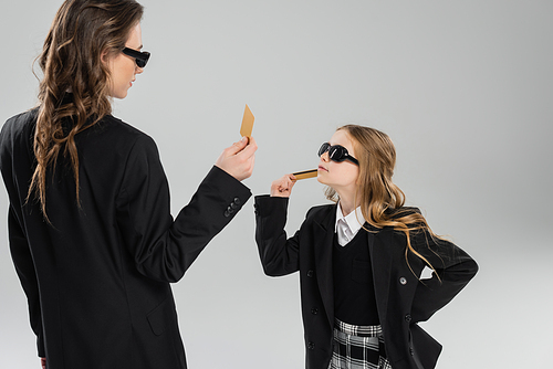 mother and daughter in sunglasses, businesswoman in suit and schoolgirl in uniform holding credit cards and looking at each other on grey background, modern parenting, money management