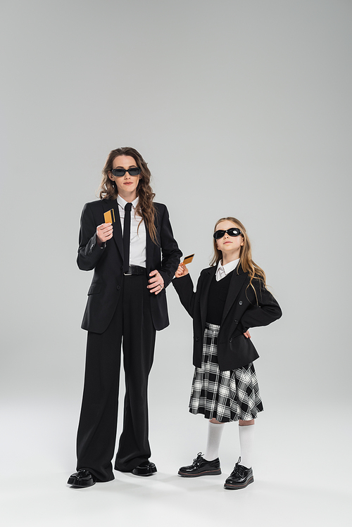 budgeting, money management, mother and daughter in sunglasses, businesswoman in suit and schoolgirl in uniform holding credit cards on grey background, modern parenting, financial learning