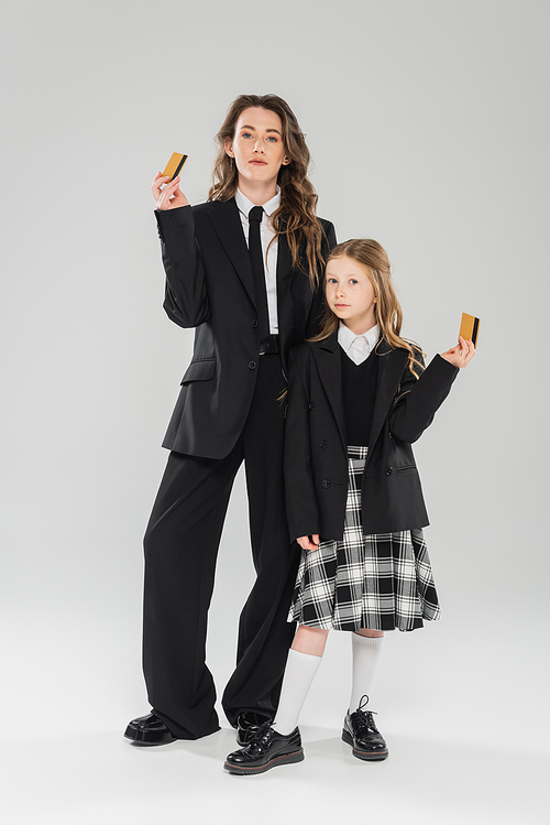 trendy mother and daughter, businesswoman in suit and schoolgirl in uniform holding credit cards on grey background, modern parenting, financial learning, budgeting, money management