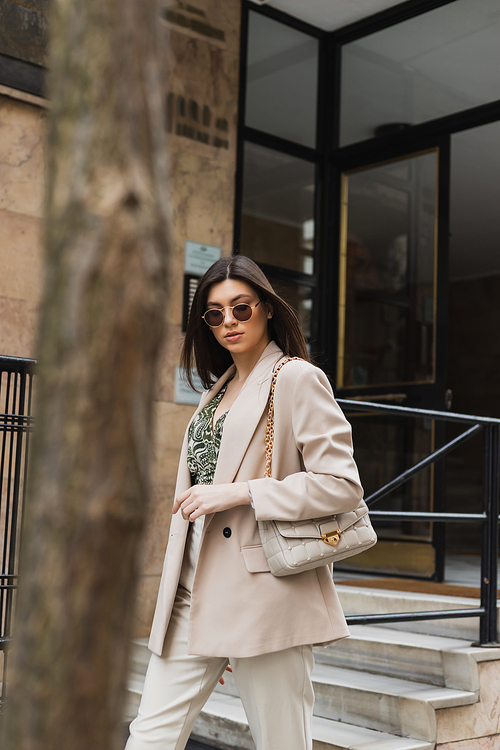 young brunette woman in stylish sunglasses and trendy outfit with white pants and beige blazer walking with handbag near modern building and blurred tree trunk on street in Istanbul,stock image