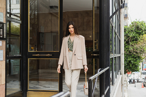 brunette and young woman in trendy outfit with white pants, blouse and beige blazer walking out of modern building while holding handbag with chain strap on street in Istanbul,stock image