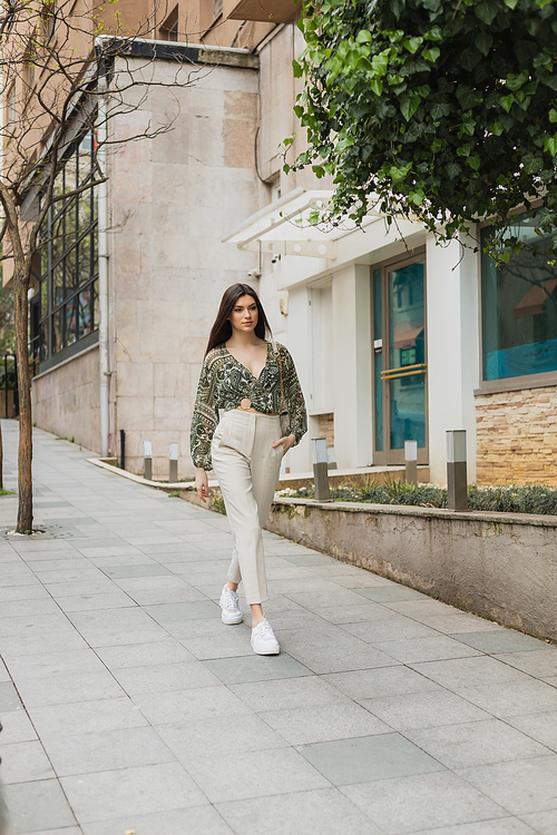 young woman with long hair in trendy outfit with beige pants, cropped blouse and handbag with chain strap walking with hand in pocket near modern building and green tree on street in Istanbul,stock image