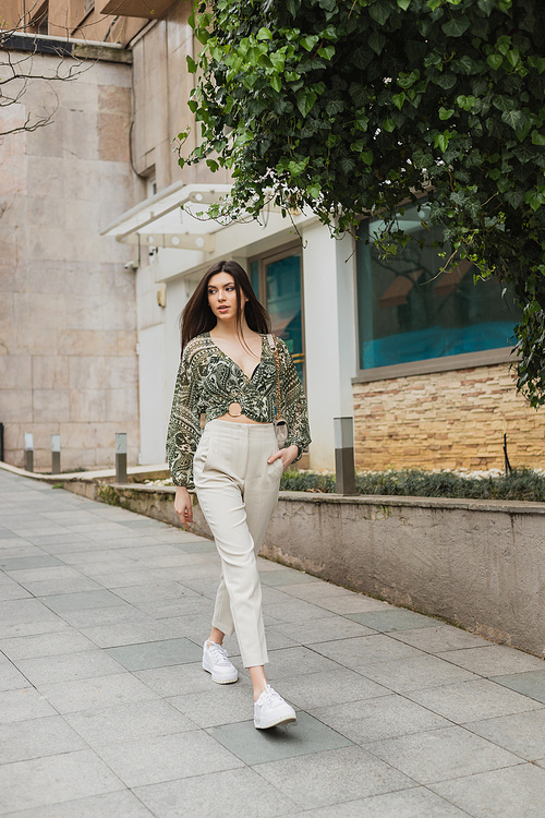 fashionable woman with long hair in trendy outfit with beige pants, cropped blouse and handbag on chain strap walking with hand in pocket near modern building and green tree on street in Istanbul,stock image