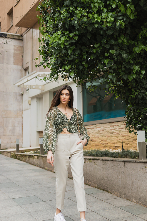 chic woman with long hair in trendy outfit with beige pants, cropped blouse and handbag with chain strap walking with hand in pocket near modern building and green tree on urban street in Istanbul,stock image