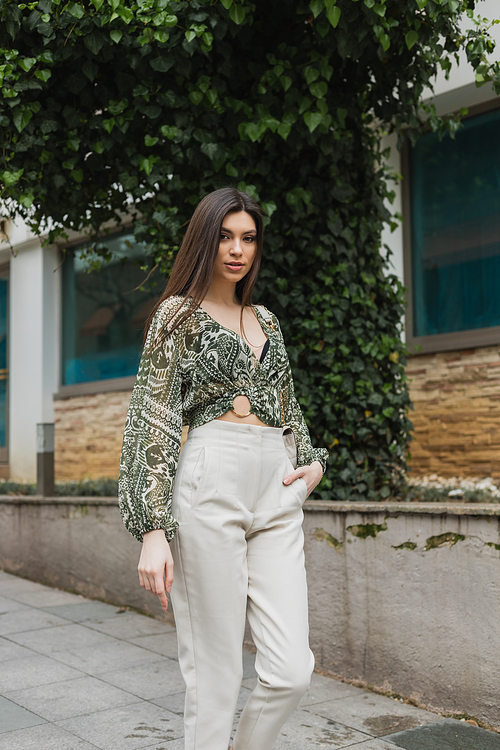 chic woman with long hair in trendy outfit with beige pants, cropped blouse and handbag with chain strap walking with hand in pocket near building and green tree on street in Istanbul,stock image