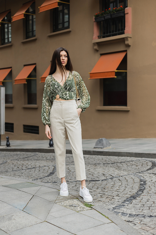 stylish woman with brunette long hair in trendy outfit with beige pants, cropped blouse and handbag with chain strap standing with hand in pocket near blurred building on urban street in Istanbul,stock image