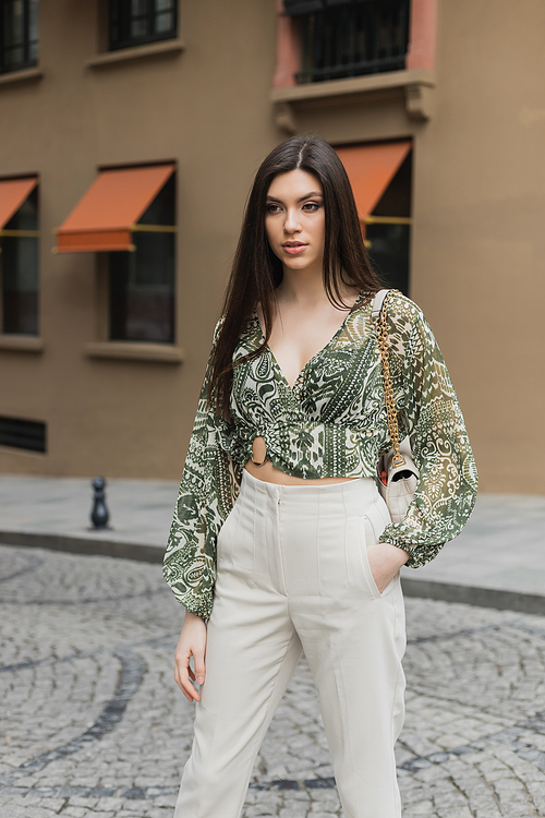 young woman with brunette long hair in beige pants, cropped blouse and handbag with chain strap standing with hand in pocket near blurred building on urban street in Istanbul,stock image