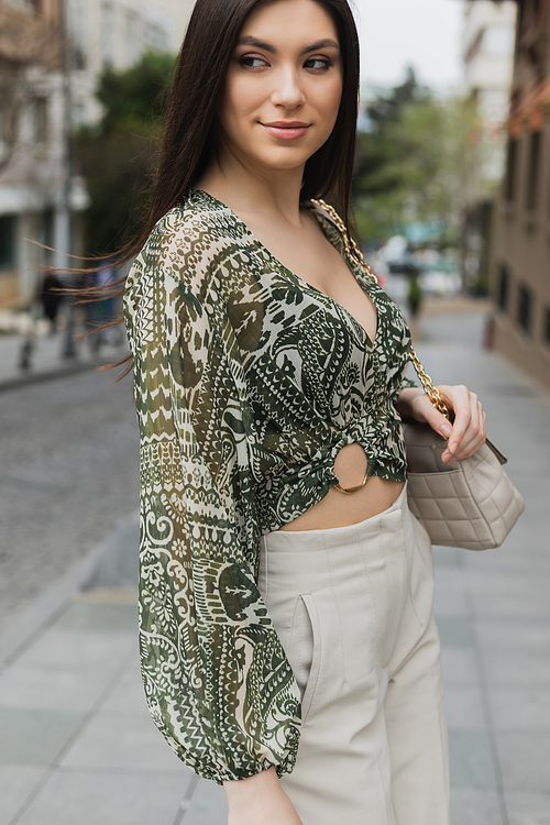 stylish woman with brunette long hair in trendy outfit with beige pants, cropped blouse and handbag with chain strap standing and smiling on urban street in Istanbul,stock image