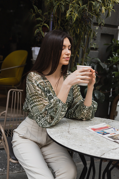 alluring woman with long brunette hair and makeup sitting on chair near round bistro table with newspaper and holding paper cup with coffee near blurred plants on terrace of cafe in Istanbul,stock image