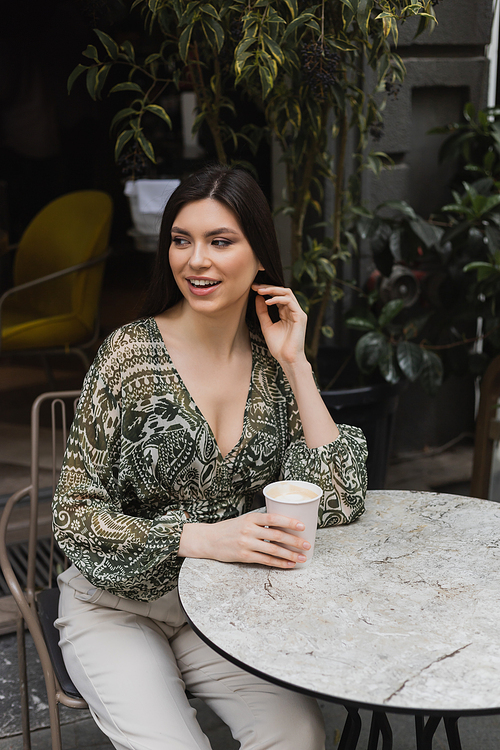 cheerful woman with long brunette hair and makeup sitting on chair near round bistro table and holding paper cup with coffee near blurred plants on terrace of cafe in Istanbul,stock image