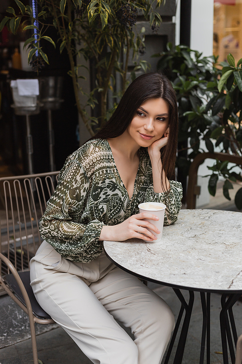 dreamy woman with long brunette hair and makeup sitting on chair near round bistro table and holding paper cup with coffee near blurred plants on terrace of cafe in Istanbul,stock image