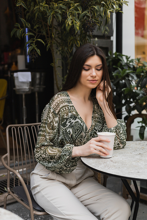 young woman with long brunette hair and makeup sitting on chair near round bistro table and looking at coffee in paper cup near blurred plants on terrace of cafe in Istanbul,stock image