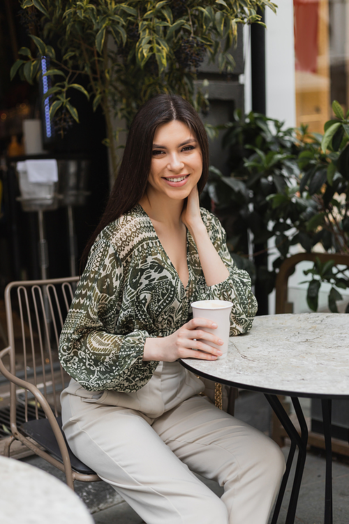 joyous young woman with long brunette hair and makeup sitting on chair near round bistro table and holding coffee in paper cup and smiling near blurred plants on terrace of cafe in Istanbul,stock image