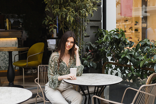 smiling woman with long brunette hair and makeup sitting on chair near round bistro table and holding coffee in paper cup near blurred plants on terrace of cafe in Istanbul,stock image