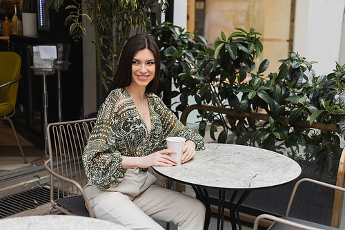 charming young woman with long brunette hair sitting on chair near round bistro table and holding coffee in paper cup while looking away and smiling near blurred plants on terrace of cafe in Istanbul,stock image