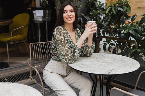 charming woman with long hair sitting on chair near round bistro table and holding paper cup with drink while looking away and smiling near blurred plants on terrace of cafe in Istanbul,stock image